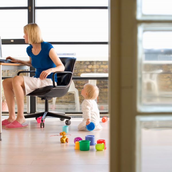 Young woman working on a computer in her home office with her baby girl on the floor playing with toys and looking up at her mother